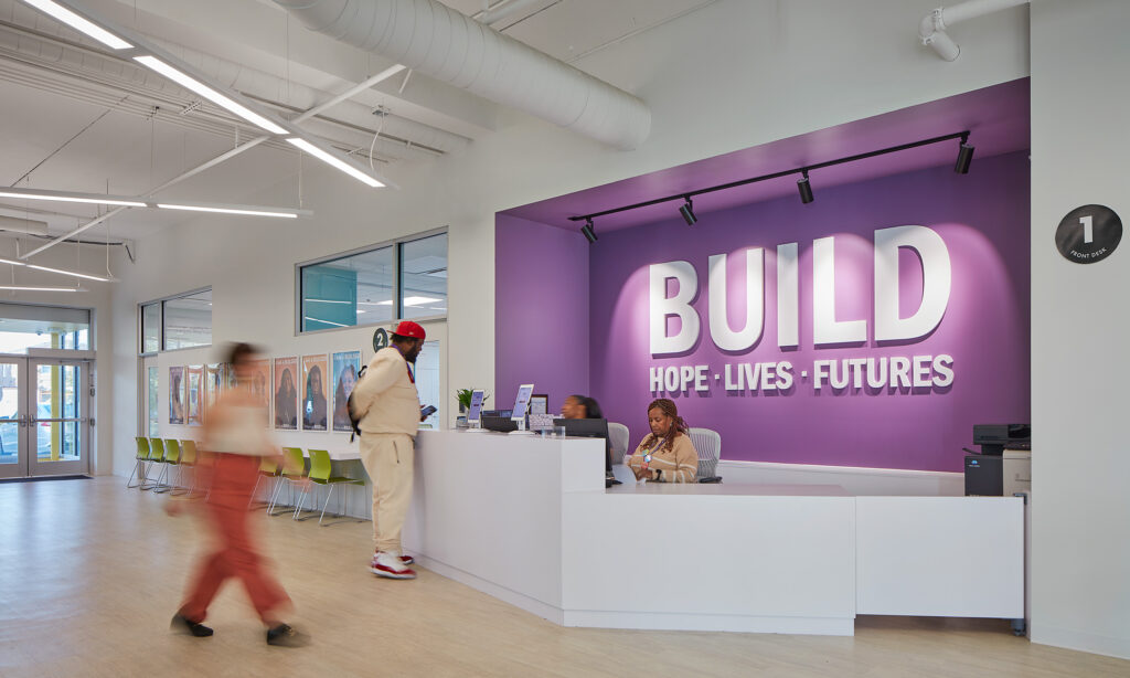Rendering of BUILD Chicago lobby. Two people walk in the lobby, while two staff members sit behind a reception desk with a purple wall behind them, with white text on the wall that reads "BUILD: Hope • Lives • Futures".